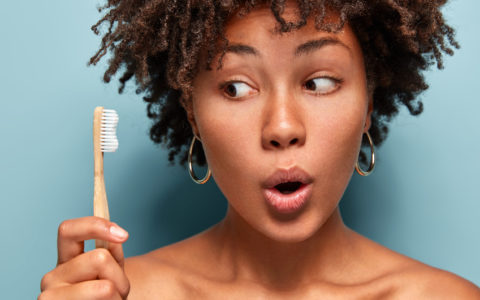 Nahaufnahme einer schockierten schönen Frau schaut auf eine hölzerne Zahnbürste, wundert sich über die brillante Wirkung der Zahnpasta, öffnet den Mund vor Überraschung, führt einen gesunden Lebensstil. Morgenstunde und tägliche Gewohnheiten