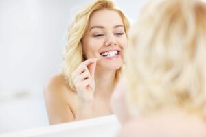 Blonde Frau steht vor Spiegel und verwendet Zahnseide